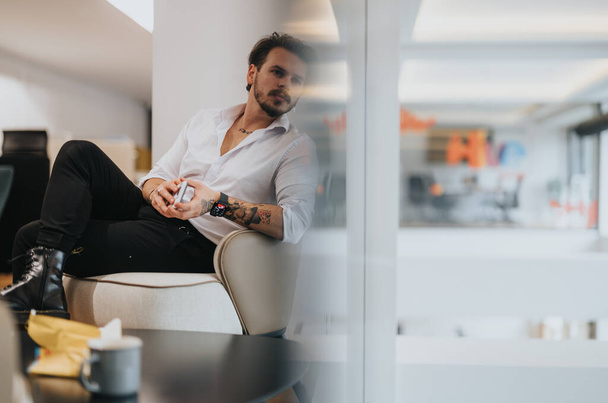 Huomaavainen tatuoitu mies raikkaassa valkoisessa paidassa lounge tyylikkäässä toimistotilassa, huokuu luottamusta ja rentoa eleganssia.. - Valokuva, kuva