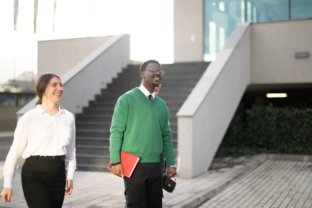 Δύο χαρούμενοι επαγγελματίες επιχειρηματίες που περπατούν μαζί έξω από ένα σύγχρονο γραφείο, συμμετέχοντας σε μια φιλική συζήτηση. - Φωτογραφία, εικόνα