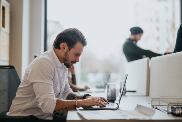 Konzentrierte männliche Person im weißen Hemd arbeitet gewissenhaft an einem Laptop in einem gut beleuchteten modernen Büroumfeld und verkörpert Produktivität. - Foto, Bild