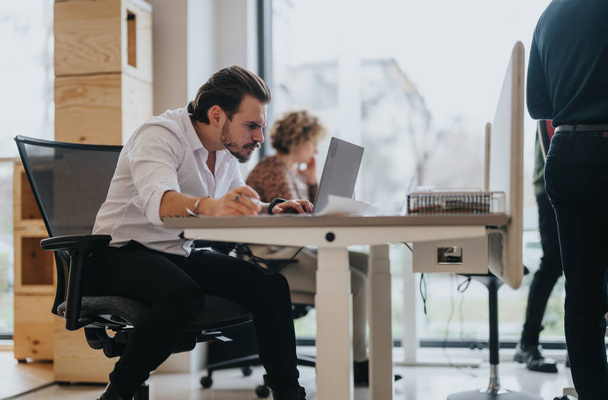 Un hombre diligente con una camisa blanca absorto en su trabajo en un ordenador portátil en un espacio de oficina contemporáneo con colegas en el fondo. - Foto, Imagen