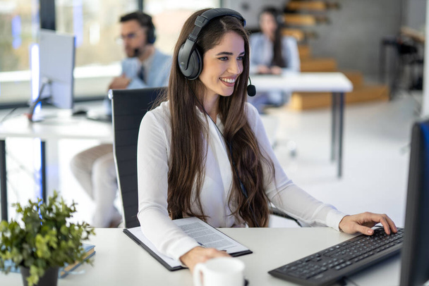 Vrouwelijke customer support agent in headset met microfoon praten met de klant en met behulp van de computer op kantoor - Foto, afbeelding