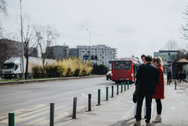 Tre individui in piedi ad una fermata dell'autobus, impegnati in una conversazione, con un autobus urbano e edifici sullo sfondo. - Foto, immagini