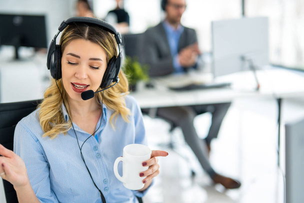 Шаріатська блондинка в гарнітурі і тримає чашку кави, розмовляючи з клієнтом під час онлайн телефонного дзвінка в службі підтримки helpdesk. - Фото, зображення