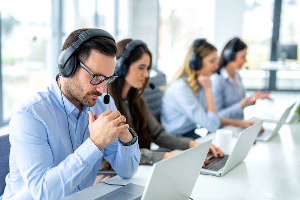 Pensive αρσενικό χειριστή εξυπηρέτησης πελατών ακούγοντας τον πελάτη μέσω ακουστικών και τη σκέψη σχετικά με τις κατάλληλες λύσεις για θέματα πελατών στο τηλεφωνικό κέντρο. Υποδοχή έννοια της ομάδας εργαζομένων. - Φωτογραφία, εικόνα