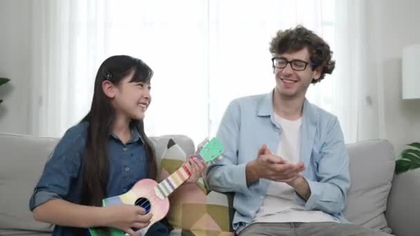 Akıllı kız oturma odasında ukulele çalarken beyaz bir babanın motivasyona ve teşviğe el çırpması. Amerikalı kız babasına gösterirken gitar öğreniyor. Aile yaratma kavramı. Pedagoji.. - Video, Çekim