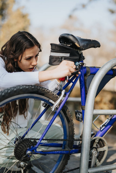 Skoncentrowana młoda kobieta zamyka swój niebieski rower na stojaku, zapewniając bezpieczeństwo podczas pobytu w parku. - Zdjęcie, obraz