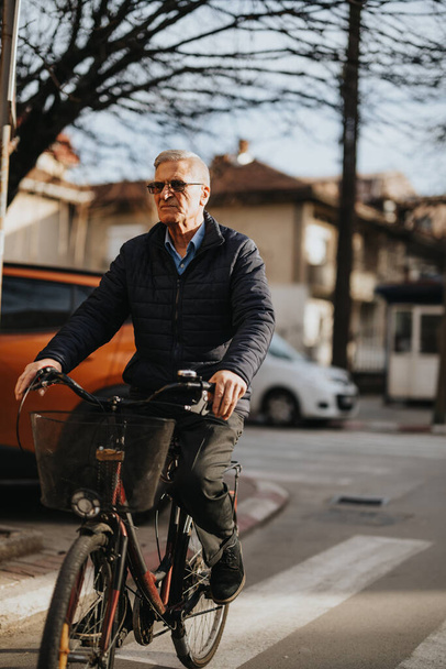 Ενεργός ηλικιωμένος άνδρας που ποδηλατεί σε δρόμο της πόλης, με έμφαση στον υγιεινό τρόπο ζωής και την αστική κινητικότητα για ηλικιωμένους. - Φωτογραφία, εικόνα
