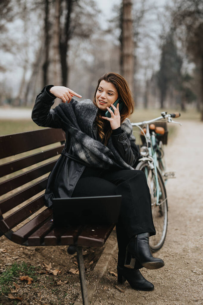 Elegantní mladá žena ve stylovém svrchním oděvu zapojena do telefonické konverzace sedí venku s kufříkem na notebook a kolo v pozadí. - Fotografie, Obrázek