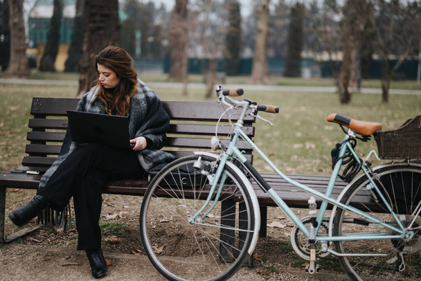 Mujer joven elegante y segura de sí misma con un atuendo elegante trabajando al aire libre en una computadora portátil, sentada en un banco junto a su bicicleta. - Foto, imagen