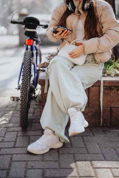 A trendy young adult rests on a city bench with her bicycle, using a smart phone and enjoying music through headphones. - Photo, Image