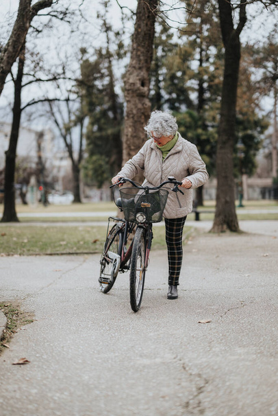 Aktiivinen vanhempi nainen kävelee polkupyörän kanssa puistopolkua pitkin ja omaksuu terveellisen elämäntavan eläkkeellä. - Valokuva, kuva