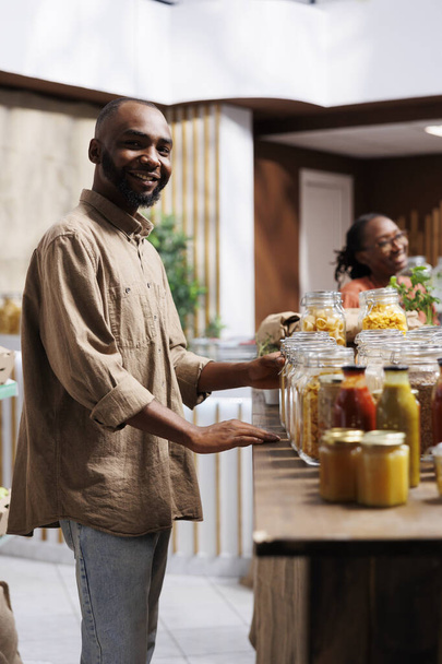 In een moderne kruidenierswinkel kijkt een gelukkige Afro-Amerikaanse man naar de camera. De planken zijn voorzien van herbruikbare verpakkingen, milieuvriendelijke producten en een selectie van items. - Foto, afbeelding