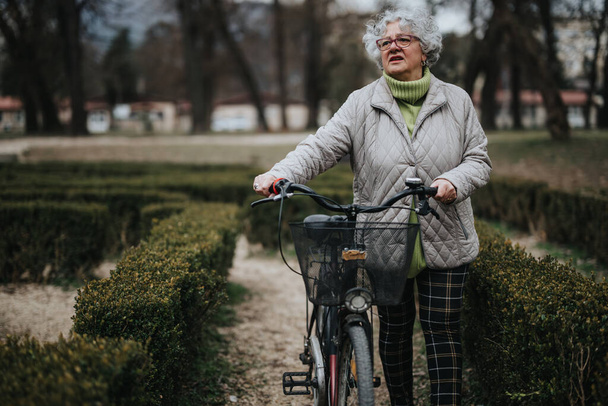 Időskorú női nyugdíjas kerékpárral, aki nyugodtan lovagol nyugodt parkban, aktív és egészséges időskort testesítve meg. - Fotó, kép