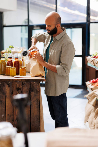 Ein Mann aus dem Nahen Osten füllt eine braune Papiertüte mit orangefarbenen Linsen. Junger Mann füllt Körner in Tüte und kauft gesunde Waren aus Mehrwegbehältern mit Bioprodukten. - Foto, Bild