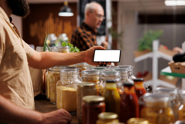 Man in zero waste shop using mockup mobile phone to analyze products. Vegan customer thoroughly checking local supermarket food items are suitable for his diet with isolated screen smartphone - Photo, Image