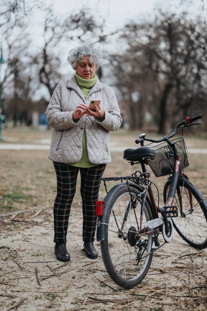 Aktive Seniorin mit schicken grauen Haaren mit Smartphone neben ihrem Fahrrad in einer ruhigen Parklandschaft. - Foto, Bild