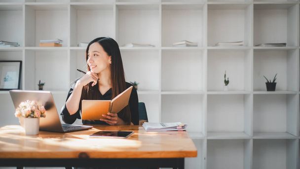 Contemplatieve zakenvrouw met een notitieboekje aan haar bureau in een minimalistisch thuiskantoor, belichaming van focus en organisatie. - Foto, afbeelding
