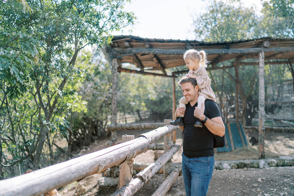Sonriente papá con niña sobre hombros de pie cerca de la cerca de madera de corral en el parque. Foto de alta calidad - Foto, Imagen