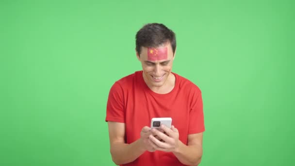 Studio video s barevným pozadím čínského fanouška s vlajkou Číny namalovanou na tváři, dívá se na svůj mobilní telefon s úsměvem a ukazuje to kameře s obrazovkou v chroma klávesy - Záběry, video
