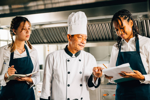 Asian senior chef teaches diverse students cooking methods in a restaurant kitchen workshop. Focusing on teamwork learning and note-taking. Professional education. Food Edocation - Fotó, kép