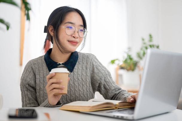 Una giovane studentessa asiatica sta sorseggiando un caffè e studiando online tramite il suo computer portatile in un caffè. persone e concetti educativi - Foto, immagini