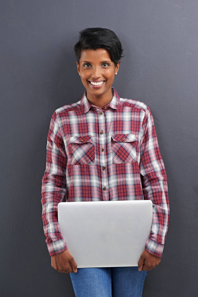 Vrolijke vrouw, portret en mode met laptop in vertrouwen op een grijze studio achtergrond. Gezicht van jonge vrouwelijke persoon, Indiase of freelancer met glimlach en computer in stijlvolle kleding op mockup ruimte. - Foto, afbeelding