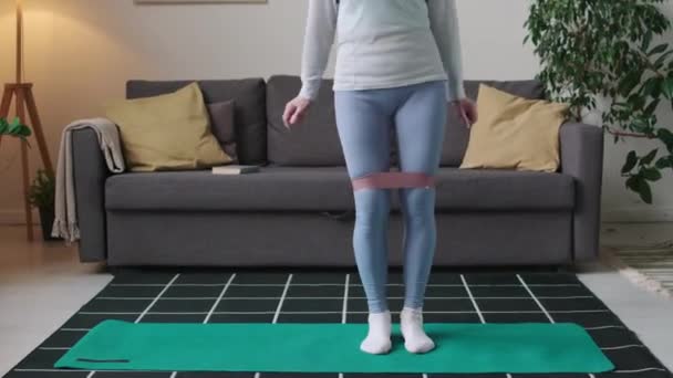Tilting up shot of Caucasian old woman doing knee banded side to side squats during domestic morning fitness training - Footage, Video