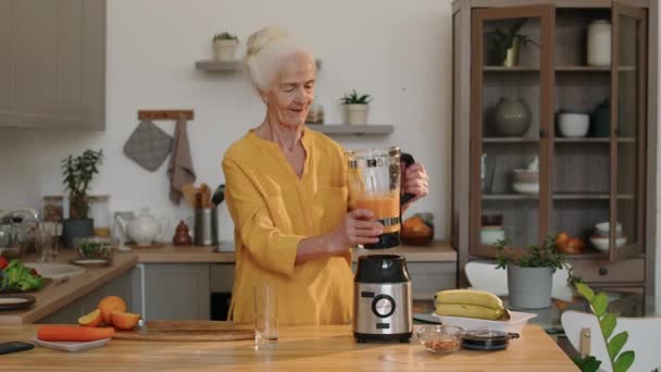 Keskipitkä kuva hyvännäköisestä valkoihoisesta vanhasta naisesta seisomassa keittiön pöydän ääressä ja kaatamassa tuoretta vitamiinismoothie-vitamiinia lasiin tehosekoittimesta. - Materiaali, video