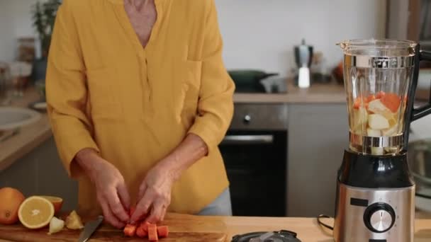Przechylając się średni strzał zdrowych białych starszych kobiet dodając rolki pokrojonej marchewki do pomarańczy i jabłek w blenderze podczas podejmowania witaminy smoothie na śniadanie w nowoczesnej kuchni - Materiał filmowy, wideo