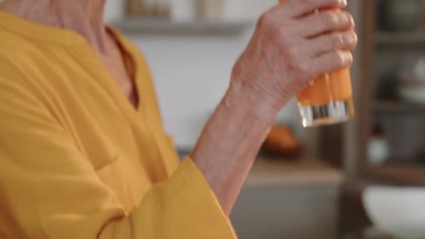 Naklápění střední detailní záběr krásné kavkazské starší ženy pití vitamín pomeranč a mrkev smoothie, stojí v moderní kuchyni v dopoledních hodinách - Záběry, video