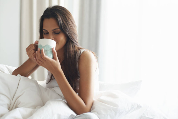 Χαμόγελο, ηρεμία και γυναίκα με καφέ στο υπνοδωμάτιο με καφεΐνη για να ξυπνήσει το πρωί στο σπίτι. Ευτυχισμένο, χαλαρώστε και άνετο νεαρό θηλυκό πρόσωπο πίνοντας κούπα καπουτσίνο, espresso ή latte στο διαμέρισμα - Φωτογραφία, εικόνα