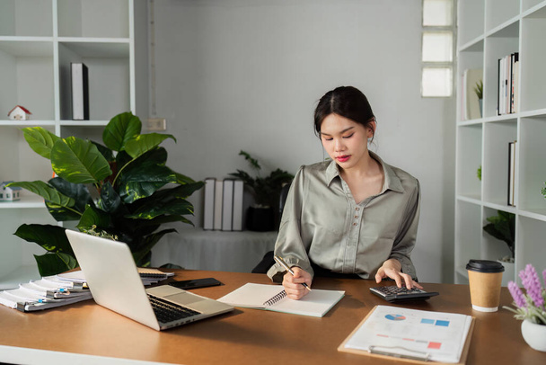 Eine Frau sitzt an einem Schreibtisch mit Taschenrechner und Stift. Sie schreibt in ein Notizbuch - Foto, Bild