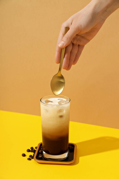 Verre avec café glacé et cuillère à la main sur table jaune sur fond beige - Photo, image