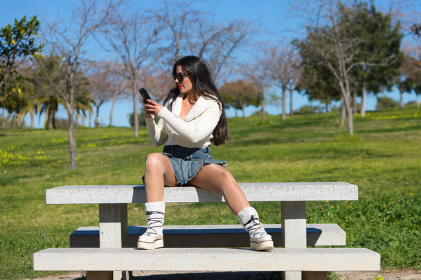 Молодая и красивая испанская брюнетка в солнечных очках сидит на скамейке в парке. Девушка небрежно одета и консультируется со своим мобильным телефоном, наслаждаясь солнечным весенним днем - Фото, изображение