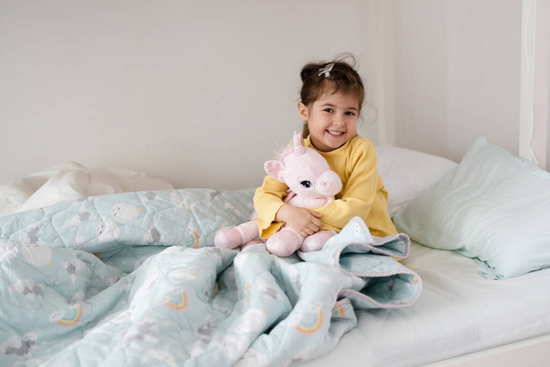 Morning awakening little child girl with her toy in bed. Happy girl in pajamas sitting on bed right after awaking. Happy family concept - Photo, Image