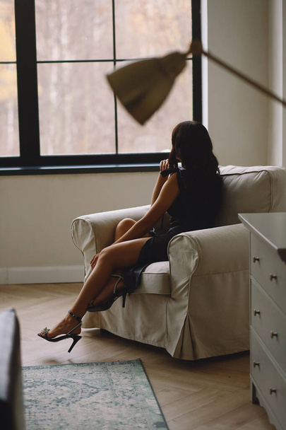 Vue arrière d'une jolie femme brune en robe noire et talons hauts avec strass assis sur la chaise dans le studio. Bon pour papier peint ou magazine d'impression - Photo, image