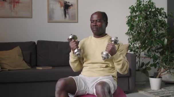 Μέτριο πλάνο του ηλικιωμένου Μαύρου άνδρα ανύψωσης αλτήρες, ενώ κάθεται σε fit μπάλα στο σύγχρονο διαμέρισμα έχουν εσωτερική προπόνηση σπορ - Πλάνα, βίντεο