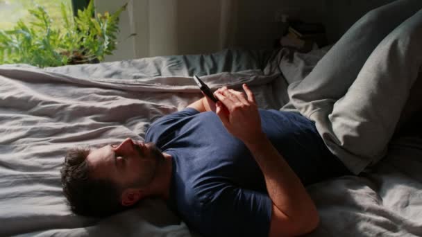 Znudzony człowiek leżący na łóżku i przeglądający telefon komórkowy - Materiał filmowy, wideo