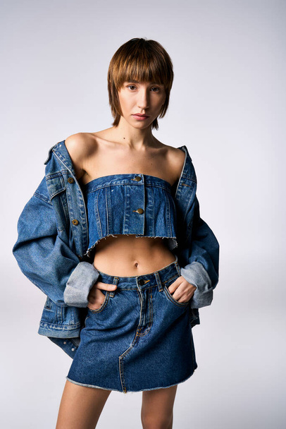 Eine junge Frau mit kurzen Haaren posiert selbstbewusst in einem trendigen Jeansrock und bauchfreiem Oberteil in einem Studio-Setting. - Foto, Bild