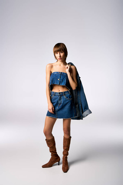 Een zelfverzekerde jonge vrouw met kort haar slaat een pose in een denim jurk en laarzen voor een fashion shoot in een studio. - Foto, afbeelding