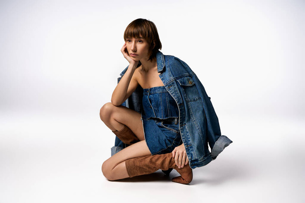 Modna młoda kobieta z krótkimi włosami elegancko siedzi na ziemi w stylowej kurtce jeansowej. - Zdjęcie, obraz