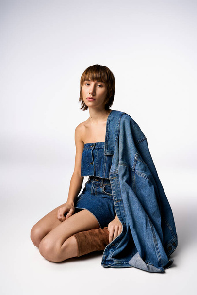 Молода жінка з коротким волоссям сидить витончено на землі в модному джинсовому платті в студійній обстановці. - Фото, зображення