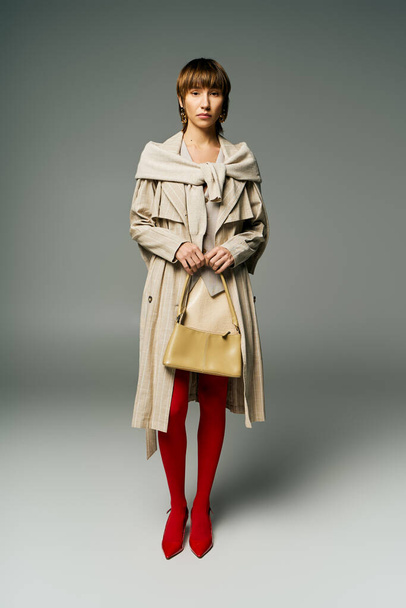 Uma jovem elegante com posses de cabelo curto em um casaco de trincheira e meias vermelhas brilhantes em um ambiente de estúdio. - Foto, Imagem