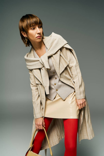 Egy elegáns fiatal nő rövid hajjal, harisnyában és kabátban, egy divatos kézitáskát tart egy stúdióban.. - Fotó, kép
