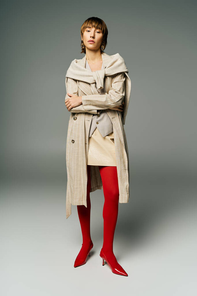 Eine junge Frau mit kurzen Haaren trägt auffällig einen Trenchcoat und leuchtend rote Strumpfhosen in einem Studio-Setting. - Foto, Bild