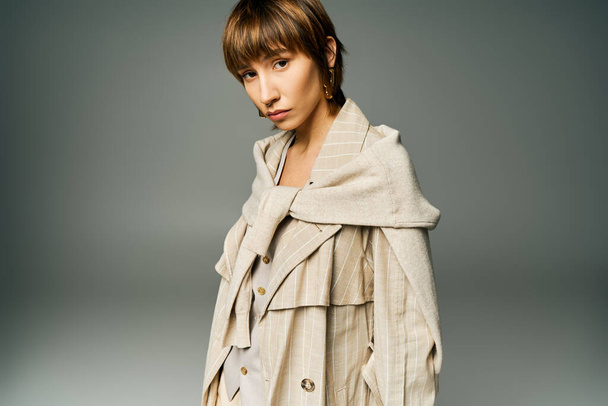 Eine stylische junge Frau mit kurzen Haaren posiert selbstbewusst im Trenchcoat für ein Porträt im Studio. - Foto, Bild