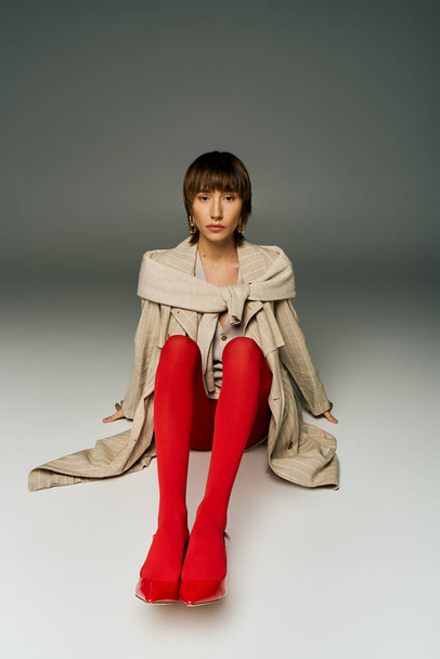 Una mujer joven con el pelo corto elegantemente se sienta en el suelo con medias rojas llamativas en un entorno de estudio. - Foto, imagen