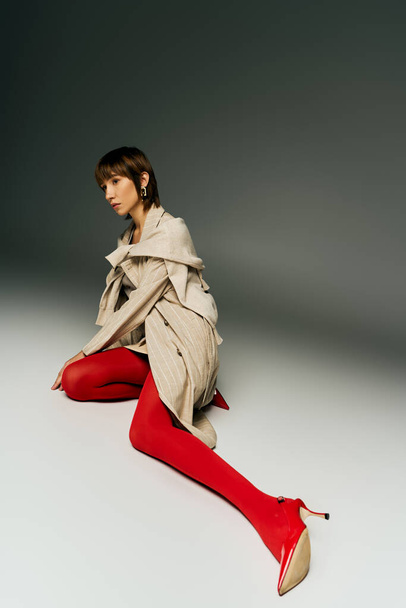 A stylish young woman with short hair poses boldly in a trench coat and vibrant red tights in a studio setting. - Fotografie, Obrázek