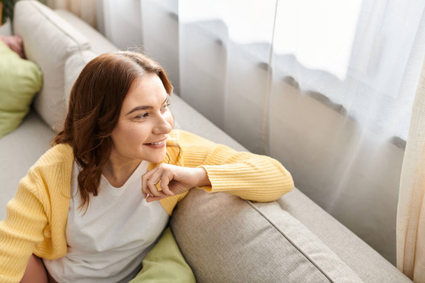 Middle aged woman in contemplation, perched on couch by a window. - Photo, Image