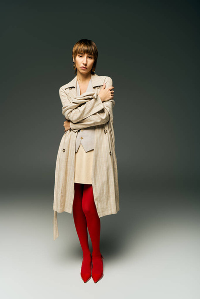 Nuori nainen lyhyet hiukset aiheuttaa studiossa yllään trenssi takki ja elinvoimainen punainen sukkahousut, tihkuu hienostuneisuutta ja tyyliä. - Valokuva, kuva